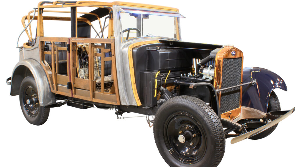 Fotografiert ist ein halb restaurierter Pkw aus der 1930er Jahren, bei dem manche Bleche noch fehlen, so dass die darunter liegende Holzkonstruktion gut zu erkennen ist.